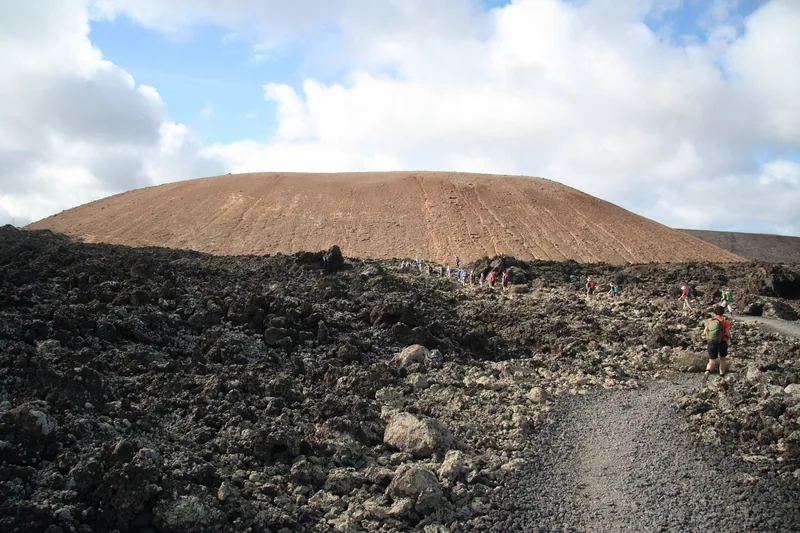 Stig genom det svarta lavafältet mot en rödbrun vulkankon på Lanzarote