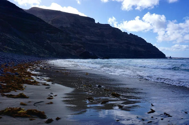 Mörk vulkansand under branta klippväggar med vågor som rullar in på Lanzarote